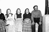 A group of people sitting together, with three women in traditional clothing and headscarves, a man in suspenders, and a baby in a lap. The women are dressed in long skirts with checkered patterns, while the man is wearing a plain shirt. The setting appears domestic with a simple backdrop.