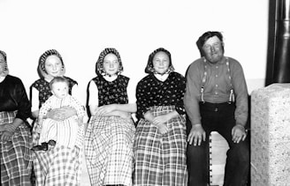 A group of people sitting together, with three women in traditional clothing and headscarves, a man in suspenders, and a baby in a lap. The women are dressed in long skirts with checkered patterns, while the man is wearing a plain shirt. The setting appears domestic with a simple backdrop.
