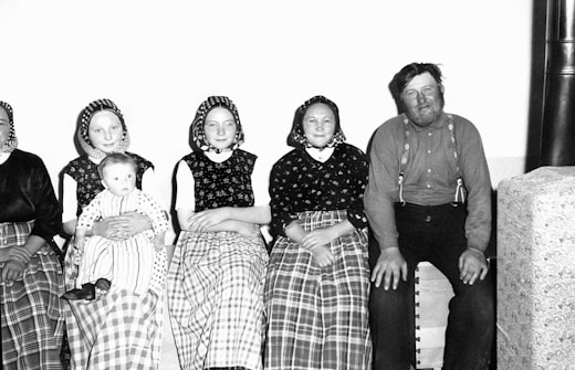 A group of people sitting together, with three women in traditional clothing and headscarves, a man in suspenders, and a baby in a lap. The women are dressed in long skirts with checkered patterns, while the man is wearing a plain shirt. The setting appears domestic with a simple backdrop.