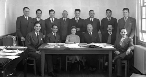 A group of thirteen people, mostly men with one woman, are gathered around a wooden table filled with papers. They are dressed in formal attire, mostly suits and ties, suggesting a professional setting. The individuals are posing for a formal group photograph with some seated and others standing behind them. The room has a simple, undecorated interior with a window on the right allowing some natural light.
