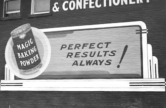 A vintage advertisement on a brick wall promoting Magic Baking Powder. The design includes a large illustration of a tin of the product, with bold text stating 'Perfect Results Always!' The advertisement uses a classic font style, with a monochromatic color scheme.