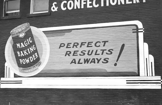 A vintage advertisement on a brick wall promoting Magic Baking Powder. The design includes a large illustration of a tin of the product, with bold text stating 'Perfect Results Always!' The advertisement uses a classic font style, with a monochromatic color scheme.