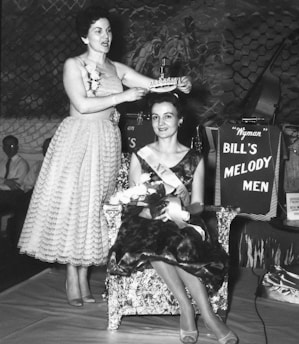 A black and white photograph captures a formal event where a woman is crowning another woman seated on an ornate chair. The seated woman is wearing a sash and holding a bouquet of flowers, likely participating in a beauty pageant. A band setup is partially visible with a sign reading 'Bill's Melody Men' in the background.