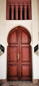 A large, ornate wooden door with an arched top set in a white façade. Above the door are decorative vertical openings with intricate designs. The door has a polished brass handle and is framed by two signs on either side, indicating directions.