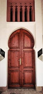 A large, ornate wooden door with an arched top set in a white façade. Above the door are decorative vertical openings with intricate designs. The door has a polished brass handle and is framed by two signs on either side, indicating directions.