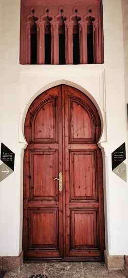 A large, ornate wooden door with an arched top set in a white façade. Above the door are decorative vertical openings with intricate designs. The door has a polished brass handle and is framed by two signs on either side, indicating directions.
