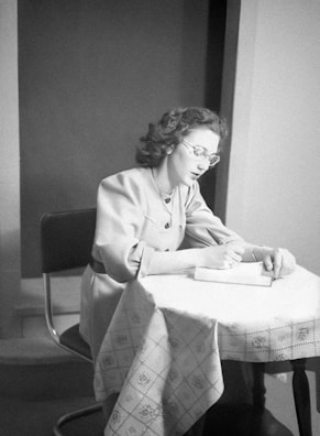 A woman with glasses and styled hair is seated at a table, writing in a notebook. She is wearing a light-colored jacket. The table has a patterned tablecloth, and the setting appears to be indoors with a neutral background.