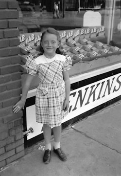 A young girl stands smiling in front of a shop display featuring stacks of FAB detergent boxes. She wears a plaid dress with puffed sleeves and has her hand resting against a brick wall. Her shoes are dark with socks, and the shop's display window is behind her.
