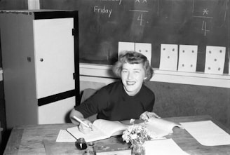 A person with short hair, dressed in a dark sweater, smiles while sitting at a desk. The desk is cluttered with open books, papers, a small bouquet of flowers in a vase, and an inkpot with a pen. Behind her, several pieces of paper with dots are pinned to a chalkboard, which also has writing on it.