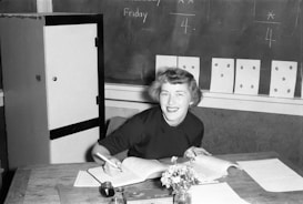 A person with short hair, dressed in a dark sweater, smiles while sitting at a desk. The desk is cluttered with open books, papers, a small bouquet of flowers in a vase, and an inkpot with a pen. Behind her, several pieces of paper with dots are pinned to a chalkboard, which also has writing on it.