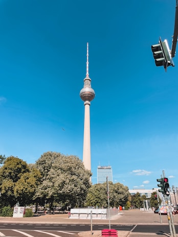 A tall telecommunications tower stands prominently against a clear blue sky. Surrounding the base are lush green trees and a street with a visible pedestrian traffic light. A building with a modern facade can be seen in the background.