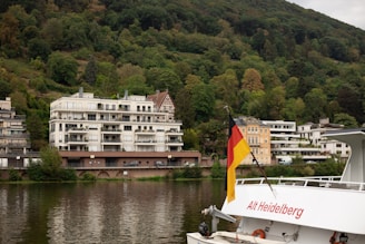 A scenic riverside view featuring modern apartment buildings situated next to a lush, green, forested hillside. A German flag is prominently displayed on a boat named 'Alt Heidelberg', which is captured in the foreground. The water reflects the surrounding foliage and structures, adding tranquility to the scene.