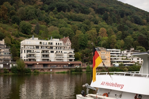 A scenic riverside view featuring modern apartment buildings situated next to a lush, green, forested hillside. A German flag is prominently displayed on a boat named 'Alt Heidelberg', which is captured in the foreground. The water reflects the surrounding foliage and structures, adding tranquility to the scene.