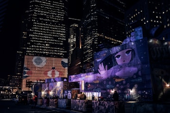 Colorful graffiti and street art panels light up a street scene at night, surrounded by towering skyscrapers. The artwork features a large anime-style character with expressive eyes, adding a whimsical contrast to the urban environment.