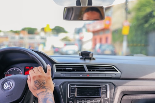 The interior of a car is visible with a person holding the steering wheel. A reflection of the driver's face appears in the rearview mirror. A tattoo is noticeable on the person's hand. The dashboard features a digital screen and control buttons. Through the windshield, an urban setting with blurred buildings and cars is seen.