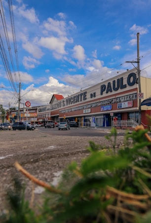 A large commercial building with signage for a store or supermarket called 'S. Vicente de Paulo'. Several parked cars are visible in front, along with people walking on the sidewalk. The sky is a bright blue with scattered clouds, and there are overhead power lines lining the street.