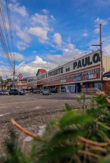 A large commercial building with signage for a store or supermarket called 'S. Vicente de Paulo'. Several parked cars are visible in front, along with people walking on the sidewalk. The sky is a bright blue with scattered clouds, and there are overhead power lines lining the street.