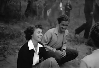 A black and white photograph of two people sitting on the ground, both smiling and appearing to be engaged in a casual conversation. The setting seems to be outdoors with blurred figures in the background, suggesting a social gathering or event.
