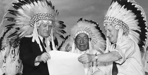 A black and white photograph featuring three men wearing traditional Native American headdresses. They are gathered closely together, examining a large piece of paper. The men are dressed in suits and ties, indicating a formal event or gathering.