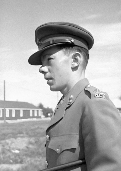 A person is dressed in a military uniform, featuring a cap and a jacket adorned with various insignia and badges. The background shows a building and a clear sky.
