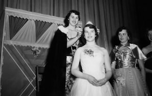 A group of women dressed in formal evening gowns are gathered in what appears to be a celebratory setting. One woman wearing a cape appears to crown another woman who is seated and wearing a strapless gown with floral embellishments. Two other women stand beside them, also in formal dresses. The background has draped curtains and decorative elements.