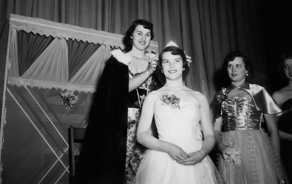 A group of women dressed in formal evening gowns are gathered in what appears to be a celebratory setting. One woman wearing a cape appears to crown another woman who is seated and wearing a strapless gown with floral embellishments. Two other women stand beside them, also in formal dresses. The background has draped curtains and decorative elements.