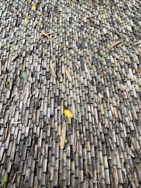 A textured surface made up of numerous small, rectangular wooden tiles. The tiles exhibit a weathered appearance with variations in color from gray to dark brown. Scattered dried leaves and debris rest on the surface, adding a natural element to the composition.