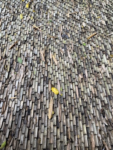 A textured surface made up of numerous small, rectangular wooden tiles. The tiles exhibit a weathered appearance with variations in color from gray to dark brown. Scattered dried leaves and debris rest on the surface, adding a natural element to the composition.