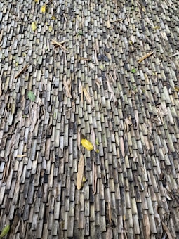 A textured surface made up of numerous small, rectangular wooden tiles. The tiles exhibit a weathered appearance with variations in color from gray to dark brown. Scattered dried leaves and debris rest on the surface, adding a natural element to the composition.