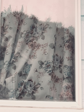 A floral curtain with a vintage design hangs behind a window. The curtain features intricate patterns of flowers and leaves in muted tones, adding a touch of elegance and nostalgia. The window frame is white, and the glass reflects subtle outdoor elements.