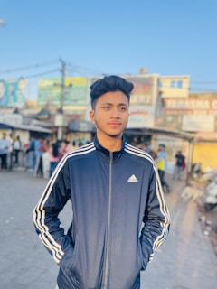 A young person is standing outdoors wearing a dark blue Adidas jacket with white stripes. In the background, there is a bustling street scene with people, shops, and various signs on buildings. The overall atmosphere is casual and lively with a clear blue sky.