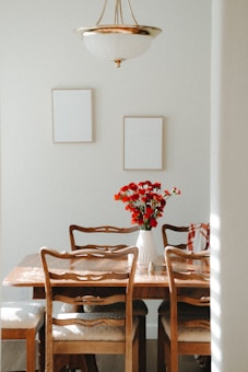 A cozy dining room setting featuring a wooden table surrounded by four chairs with upholstered seats. A white vase with vibrant red flowers is centrally placed on the table. Two blank picture frames hang on the light-colored wall, and an elegant chandelier with metallic accents is suspended above the table. Light from an unseen window casts gentle shadows across the room.