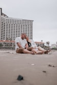 A couple sits closely on a sandy beach in a relaxed and affectionate pose. In the background, there is a high-rise hotel building with numerous balconies. The sky appears overcast, and the atmosphere feels calm and serene.