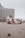 A couple sits closely on a sandy beach in a relaxed and affectionate pose. In the background, there is a high-rise hotel building with numerous balconies. The sky appears overcast, and the atmosphere feels calm and serene.