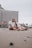 A couple sits closely on a sandy beach in a relaxed and affectionate pose. In the background, there is a high-rise hotel building with numerous balconies. The sky appears overcast, and the atmosphere feels calm and serene.