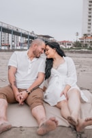 A couple sits closely on a sandy beach, sharing an intimate moment with their foreheads touching. The man is wearing a white shirt and brown shorts, while the woman is dressed in a white dress. Their expressions are warm and affectionate. Behind them, a pier extends into the horizon and residential buildings are visible.