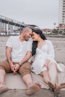 A couple sits closely on a sandy beach, sharing an intimate moment with their foreheads touching. The man is wearing a white shirt and brown shorts, while the woman is dressed in a white dress. Their expressions are warm and affectionate. Behind them, a pier extends into the horizon and residential buildings are visible.