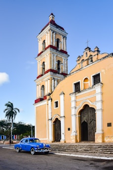 An old, colorful church with a tall bell tower is situated in a quiet town square. The walls of the church display architectural details typical of colonial design, with arches and ornate windows. A classic blue car is parked in front of the building, adding a nostalgic feel to the scene. A tall palm tree stands nearby under a clear blue sky.