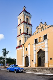 An old, colorful church with a tall bell tower is situated in a quiet town square. The walls of the church display architectural details typical of colonial design, with arches and ornate windows. A classic blue car is parked in front of the building, adding a nostalgic feel to the scene. A tall palm tree stands nearby under a clear blue sky.