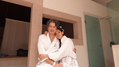 A man and a woman wearing white robes are sitting closely together, appearing relaxed and affectionate. They are in a modern room with light-colored walls and a glass shower door.