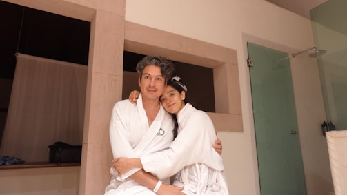 A man and a woman wearing white robes are sitting closely together, appearing relaxed and affectionate. They are in a modern room with light-colored walls and a glass shower door.