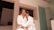 A man and a woman wearing white robes are sitting closely together, appearing relaxed and affectionate. They are in a modern room with light-colored walls and a glass shower door.