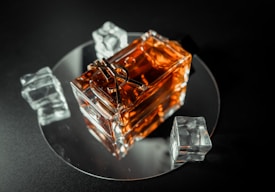 A glass bottle with a golden liquid sits on a reflective circular surface. Surrounding the bottle are three clear, faceted ice cubes. The scene is set against a dark background, highlighting the warm tones of the liquid.