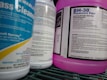 Several bottles of cleaning products are arranged close together on a shelf. Labels on the bottles provide information about the contents, warnings, and usage instructions. The prominent colors are blue and purple, with detailed text visible on the labels.
