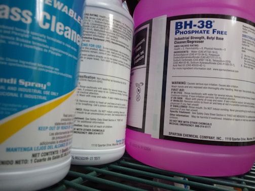 Several bottles of cleaning products are arranged close together on a shelf. Labels on the bottles provide information about the contents, warnings, and usage instructions. The prominent colors are blue and purple, with detailed text visible on the labels.