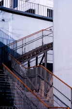 A detailed view of an outdoor metal staircase with contrasting shadows and light. The structure features multiple railings and steps, highlighted by natural sunlight. The surrounding walls are painted in a light blue shade, and there is a small wall-mounted light fixture on the upper left side.