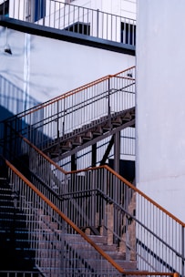 A detailed view of an outdoor metal staircase with contrasting shadows and light. The structure features multiple railings and steps, highlighted by natural sunlight. The surrounding walls are painted in a light blue shade, and there is a small wall-mounted light fixture on the upper left side.