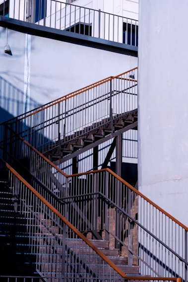 A detailed view of an outdoor metal staircase with contrasting shadows and light. The structure features multiple railings and steps, highlighted by natural sunlight. The surrounding walls are painted in a light blue shade, and there is a small wall-mounted light fixture on the upper left side.