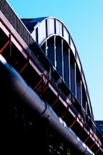 A close-up view of a large steel structure, possibly part of a bridge or industrial building, with curved beams and metal girders in shades of silver and black. The structure contrasts against a clear blue sky, and the light emphasizes the texture and rivets of the metal.