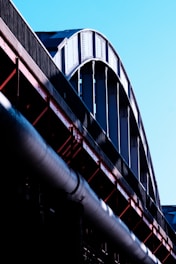 A close-up view of a large steel structure, possibly part of a bridge or industrial building, with curved beams and metal girders in shades of silver and black. The structure contrasts against a clear blue sky, and the light emphasizes the texture and rivets of the metal.